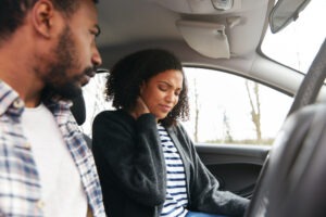 A passenger sits in a car with the driver holding her neck in pain after an accident.