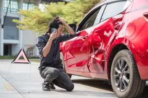 A man looks at the damage on his red sedan, wondering what to do after a car accident.